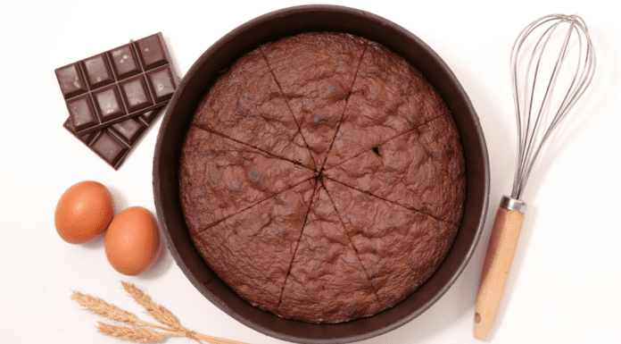 A round chocolate cake cut into 8 pieces. Two brown eggs and bars of choclate on the left with a wisk on the right side.