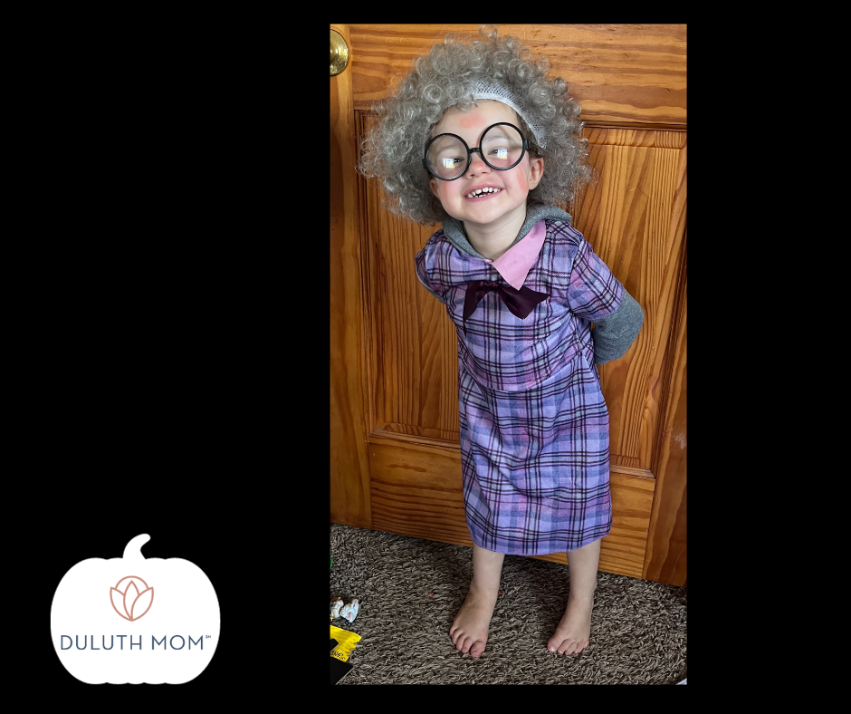 toddler dressed with a gray curly wig, large round circle black framed glasses and a purple plaid dress