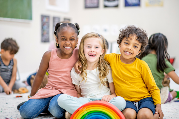 Three kids with their arms around eachother smiling with a rainbow toy set infront of them