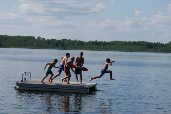 4 boys jumping off of a raft into a lake with a life guard watching them