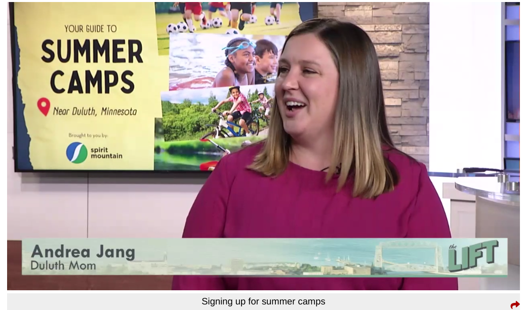 Andrea is wearing a magenta shirt sitting in front of a screen showing a yellow summer camp graphic. 